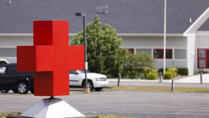 Large red cross sculpture in a parking lot, positioned in front of a white building with red-trimmed windows and surrounding greenery, indicating a healthcare or social services facility.