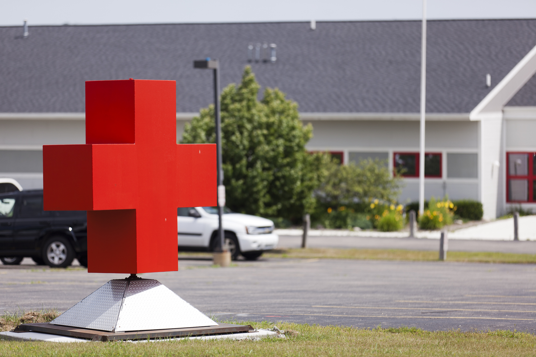 Large red cross sculpture in a parking lot, positioned in front of a white building with red-trimmed windows and surrounding greenery, indicating a healthcare or social services facility.